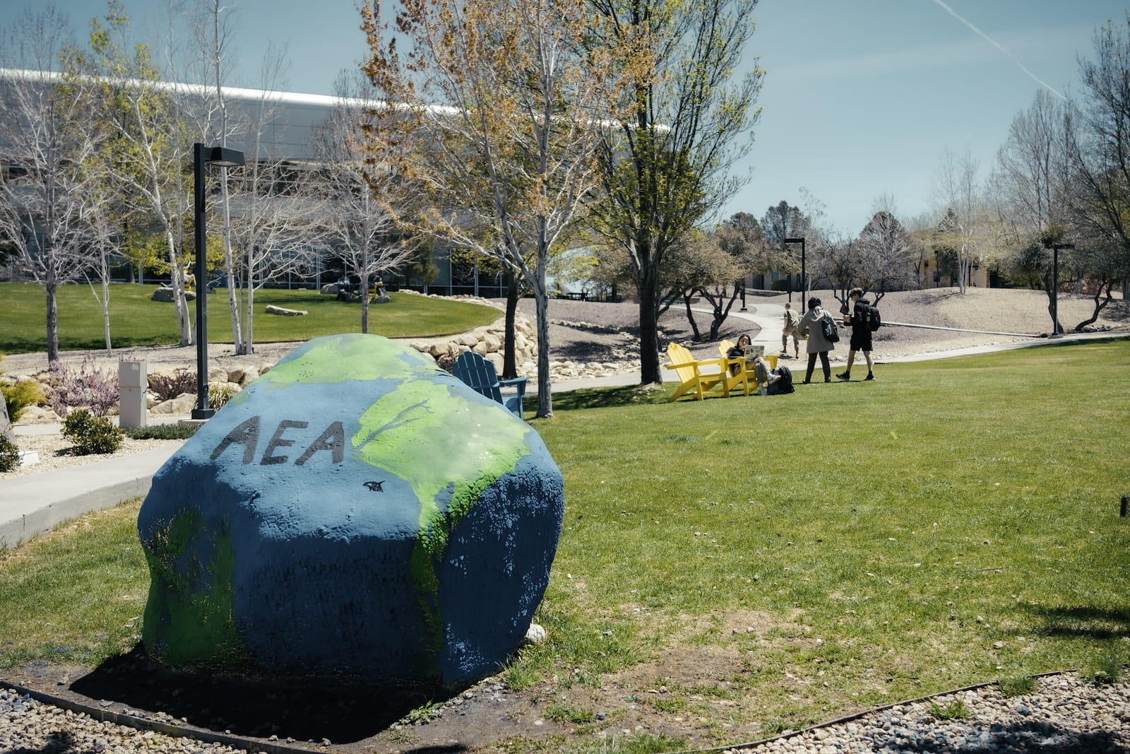 Boulder on the Prescott campus painted like a globe with the letters 'AEA' in the foreground with students walking and sitting on bright Adirondack chairs across a grassy lawn.