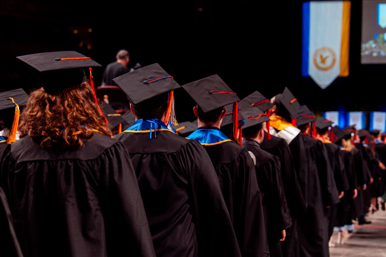 Graduates in cap and gown lined up in procession during commencement ceremony with university banner visible on stage.