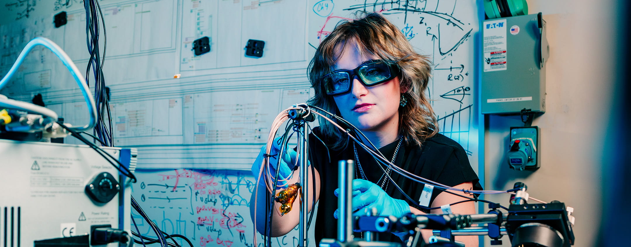 Student working with biomedical instrumentation and tubing setup in engineering lab, wearing protective glasses and gloves, with circuit diagrams and equations on whiteboard behind.