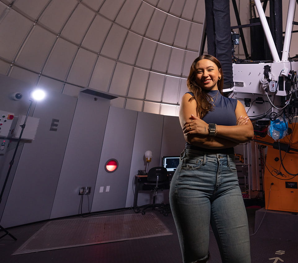Student stands inside an observatory dome beside a large telescope mount and control equipment, with curved dome panels and lighting visible in the lab environment.
