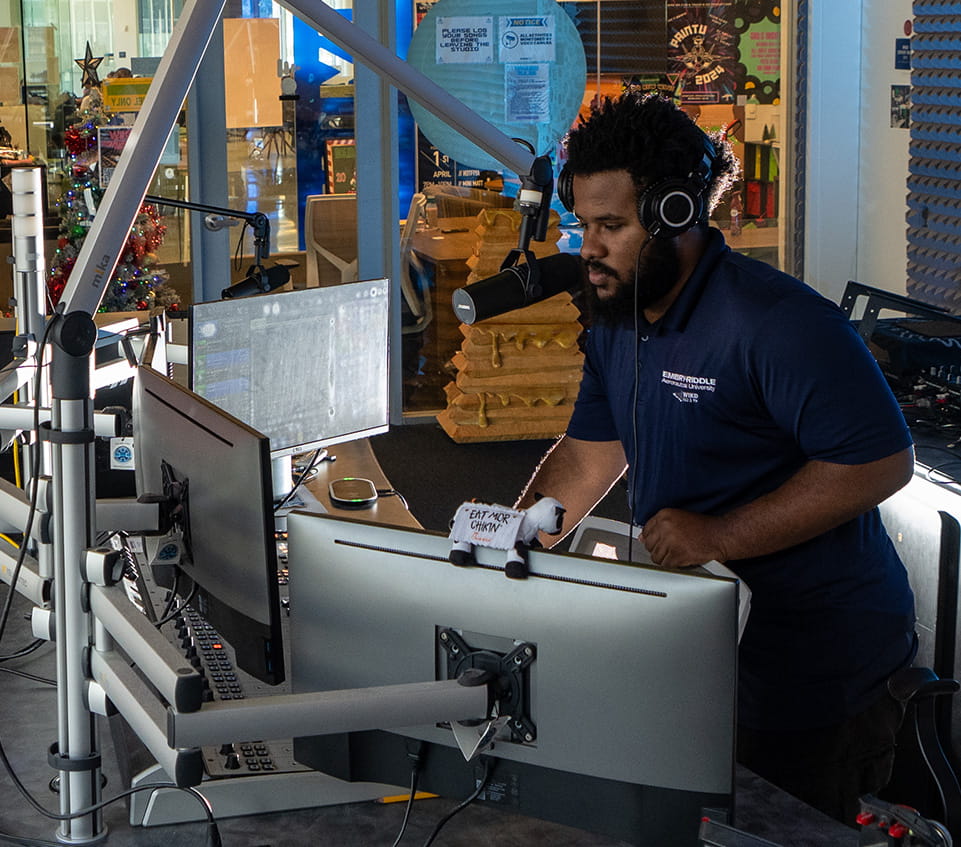 Student operates a campus radio studio, speaking into a broadcast microphone with headphones on, using a multi-monitor audio workstation with boom arms, mixer controls, and acoustic foam panels visible behind glass.