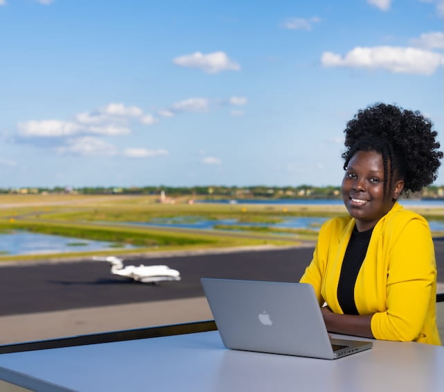 Student in a bright yellow cardigan sits at a laptop, smiling, with a bright open landscape in the background.