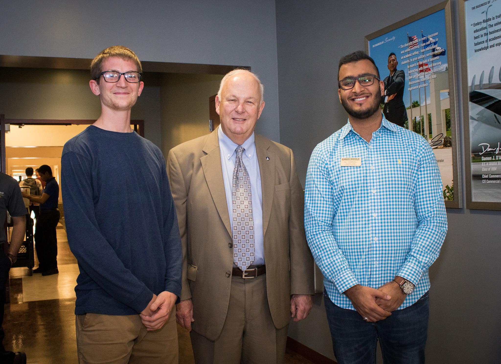 Ali Riza Karmali and a student pose with David B. O'Maley together in campus hallway with framed aerospace and alumni displays on wall.