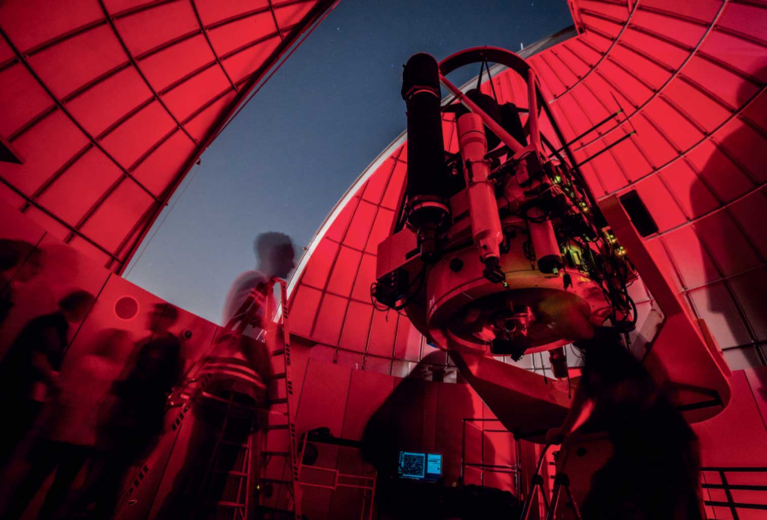 Large research telescope mounted inside observatory dome with open shutter, students moving around instrument under red light for nighttime observation.