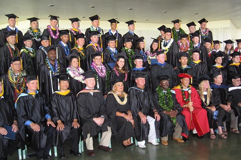 Large group of graduates and faculty in caps and gowns seated and standing on stage, academic regalia with hoods and stoles.