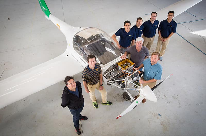 Group of students and instructor gathered around small single-engine training aircraft with cowling removed, exposed engine and propeller assembly visible inside hangar, high-angle view.