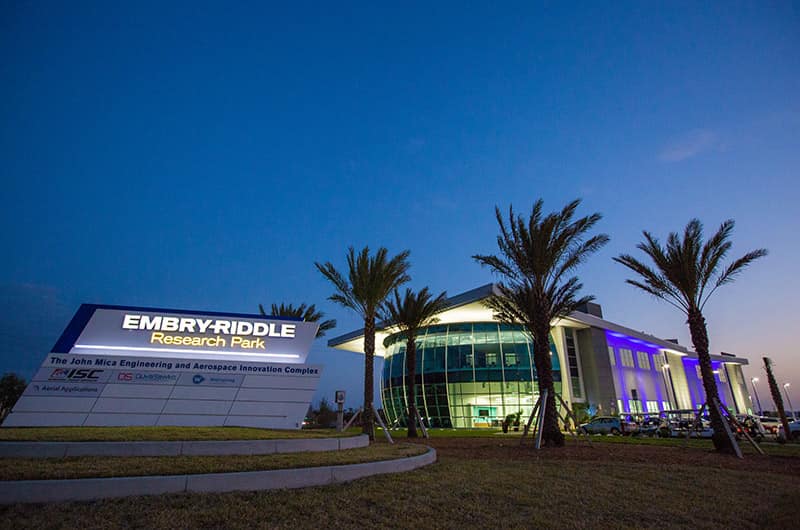 MicaPlex building with glass facade and illuminated entrance at dusk, palm trees lining walkway and campus signage visible.