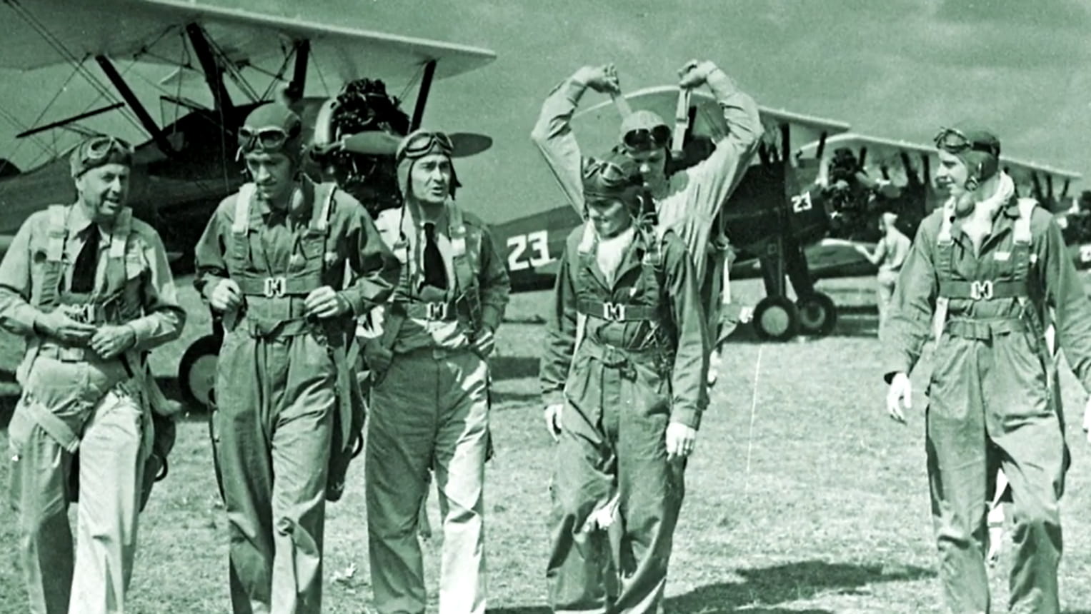World War II-era pilots in flight suits and parachute harnesses walking on airfield in front of military aircraft, in a vintage black and white photo.