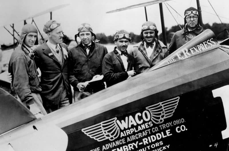 Open-cockpit 1927 biplane with 'Waco Aircraft Co.' marking on fuselage, group of early aviators and suited men gathered around and seated in cockpit, black and white photo.