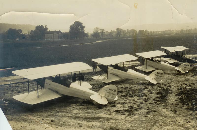 Line of early open-cockpit biplanes parked on dirt airfield with simple hangar structures in background, black and white photo.