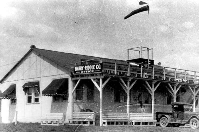 Small airfield terminal building with covered porch and observation deck, windsock above roof and early automobile parked nearby, black and white photo.