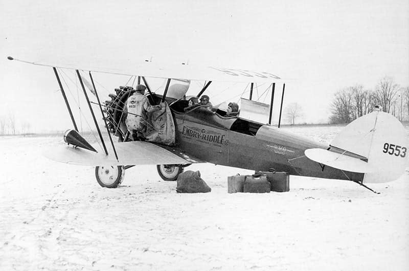 Early biplane with two pilots in the open cockpit, and a mechanic working from a wing. Suitcases are on the field near the tail.