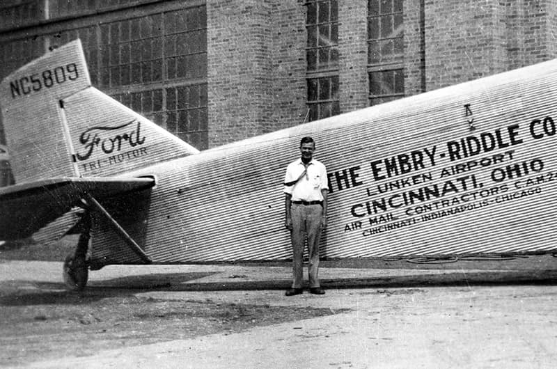 Man standing beside Ford Trimotor with 'The Embry-Riddle Co., Lunken Airport, Cincinnati, Ohio' painted on fuselage, corrugated metal tri-motor aircraft, black and white photo.