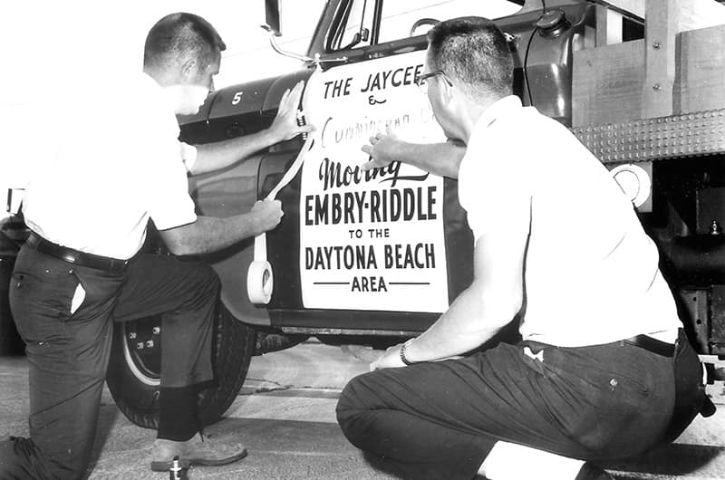 Men pasting a sign to a truck door that reads, 'The Jaycees and Cunningham moving Embry-Riddle to the Daytona Beach area.'