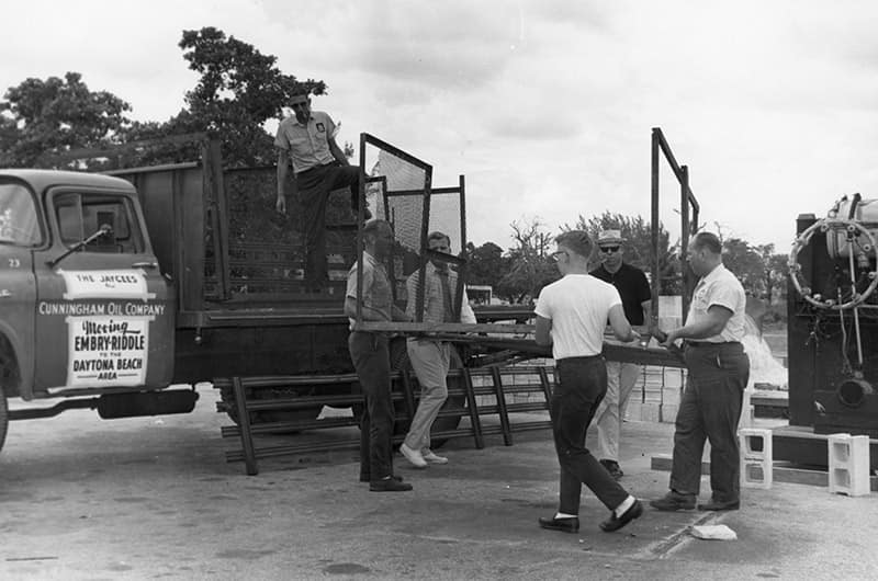 Workers loading equipment onto truck with 'Embry-Riddle Aeronautical University' signage on door, industrial yard with machinery and crates, black and white photo.