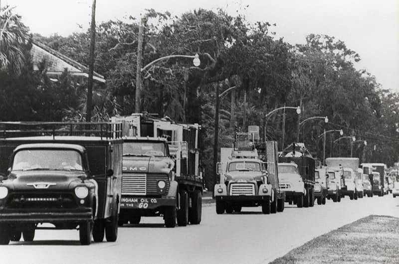 Convoy of utility trucks driving along road lined with trees, black and white photo.