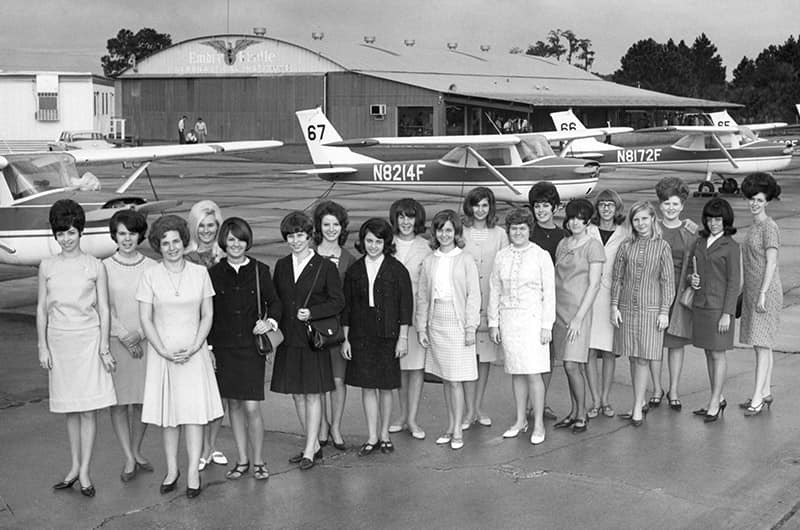 Group of women in 60s-style dresses standing on airfield in front of small training aircraft and hangar, posed lineup, black and white photo.
