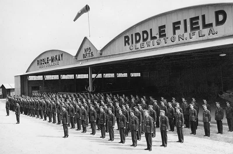 Rows of uniformed cadets standing in formation outside Riddle Field hangar with large 'RIDDLE FIELD DAYTONA FLA' signage, aircraft parked on roof above entrance, black and white image.