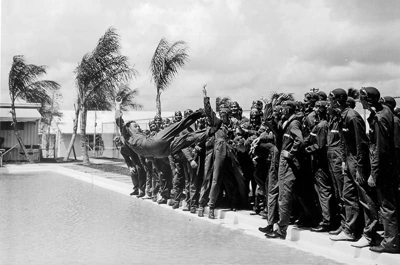 Group of cadets in uniform gathered outdoors raising right hands in unison, palm trees bending in strong wind behind them near campus buildings, black and white image.
