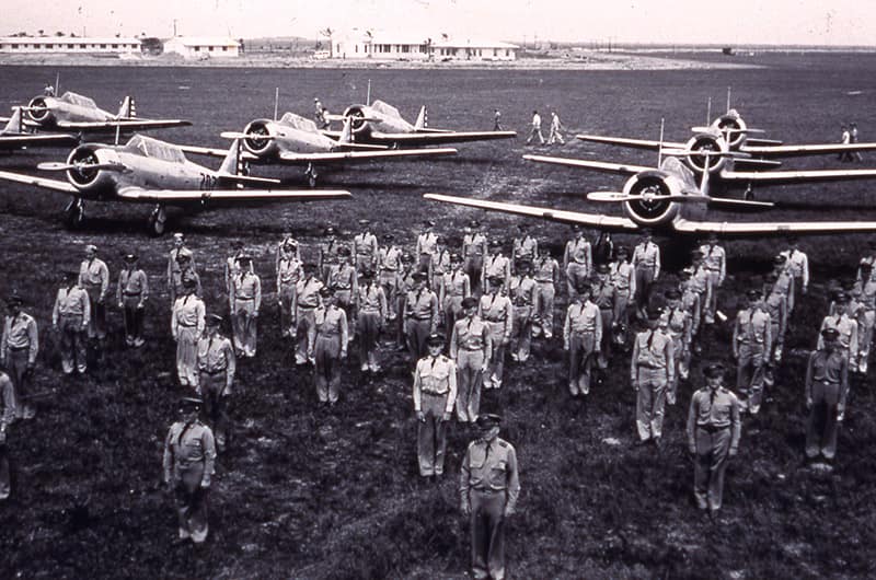 Rows of cadets standing in formation on grass in front of several single-engine training aircraft parked on runway, black and white image.