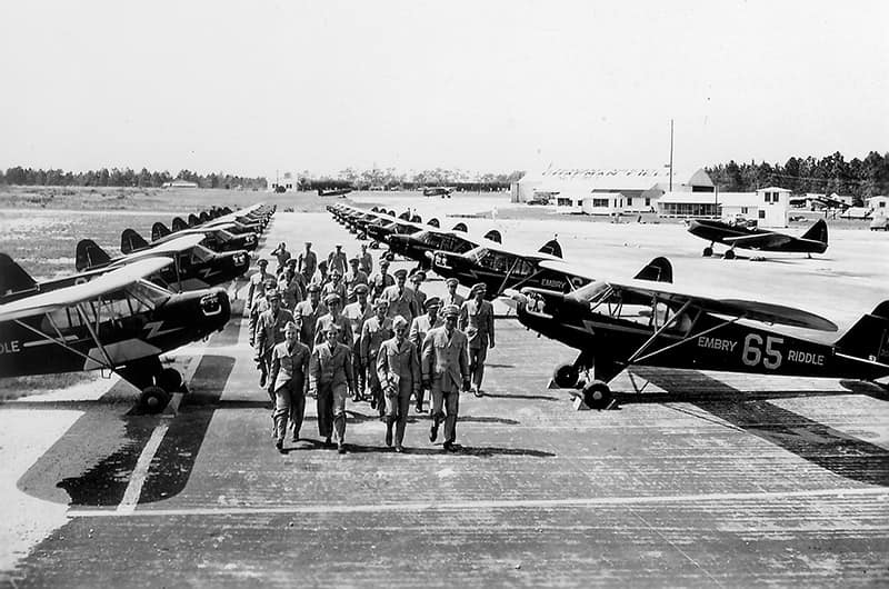 Large formation of cadets marching down runway between rows of parked single-engine training aircraft, control tower visible in distance, black and white image.