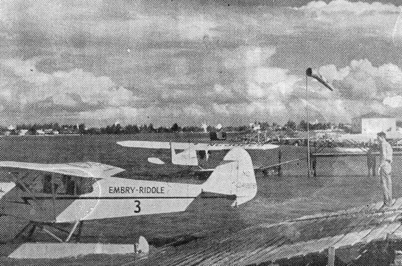 Larsen-Riddle training aircraft number 3 parked on grass airfield with another light aircraft nearby and staff standing beside runway, early campus buildings in background, black and white image.
