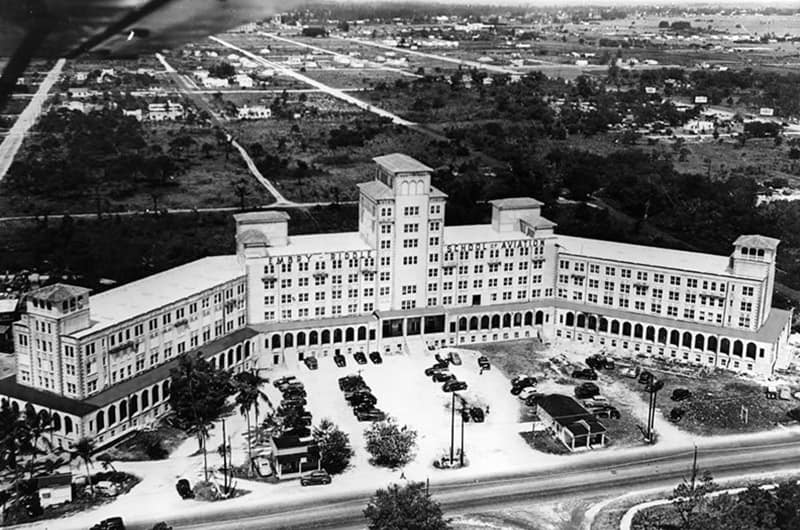 Aerial view of large Mediterranean Revival–style campus building with central tower and curved wings surrounding courtyard with palm trees, surrounding city and coastline in distance, black and white image.