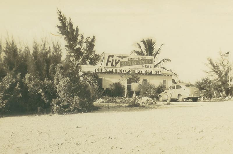 Small roadside office with 'Fly Embry-Riddle' sign above entrance, palm trees and parked cars beside sandy lot, black and white image.