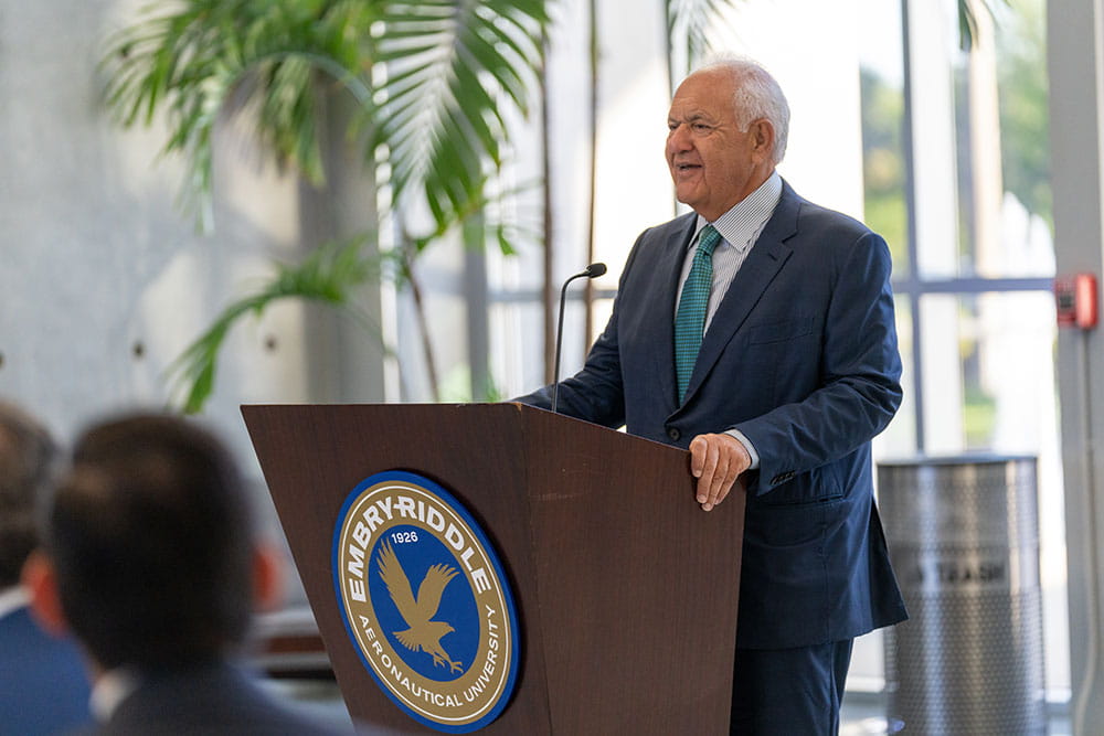 Board of Trustees chair Mori Hosseini, in a blue suit and tie, speaking at a podium with the Embry-Riddle Aeronautical University seal, addressing an audience in a bright indoor space.