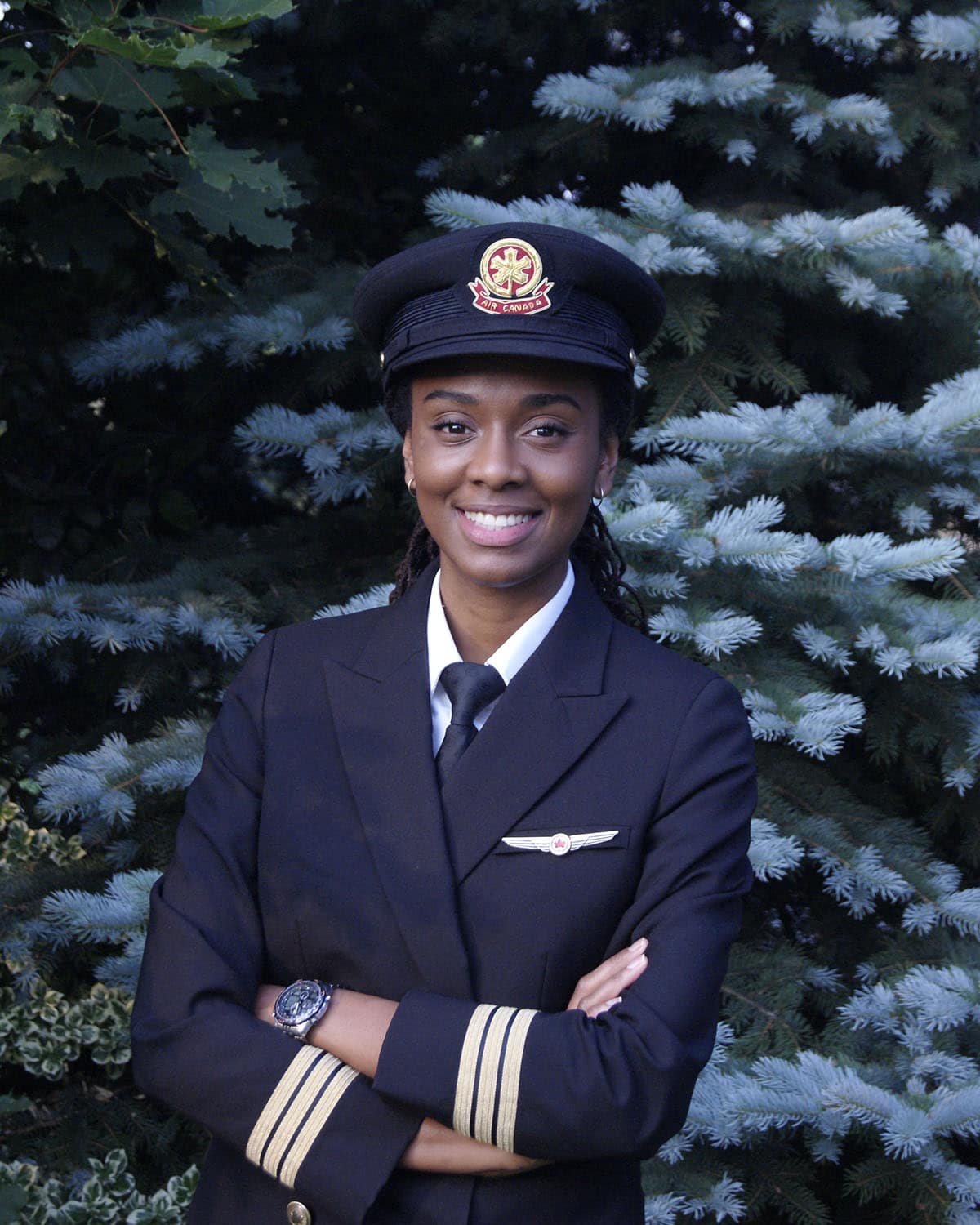 Zoey Williams in her Air Canada uniform with captain-style hat and sleeve stripes, arms crossed, standing in front of evergreen trees.