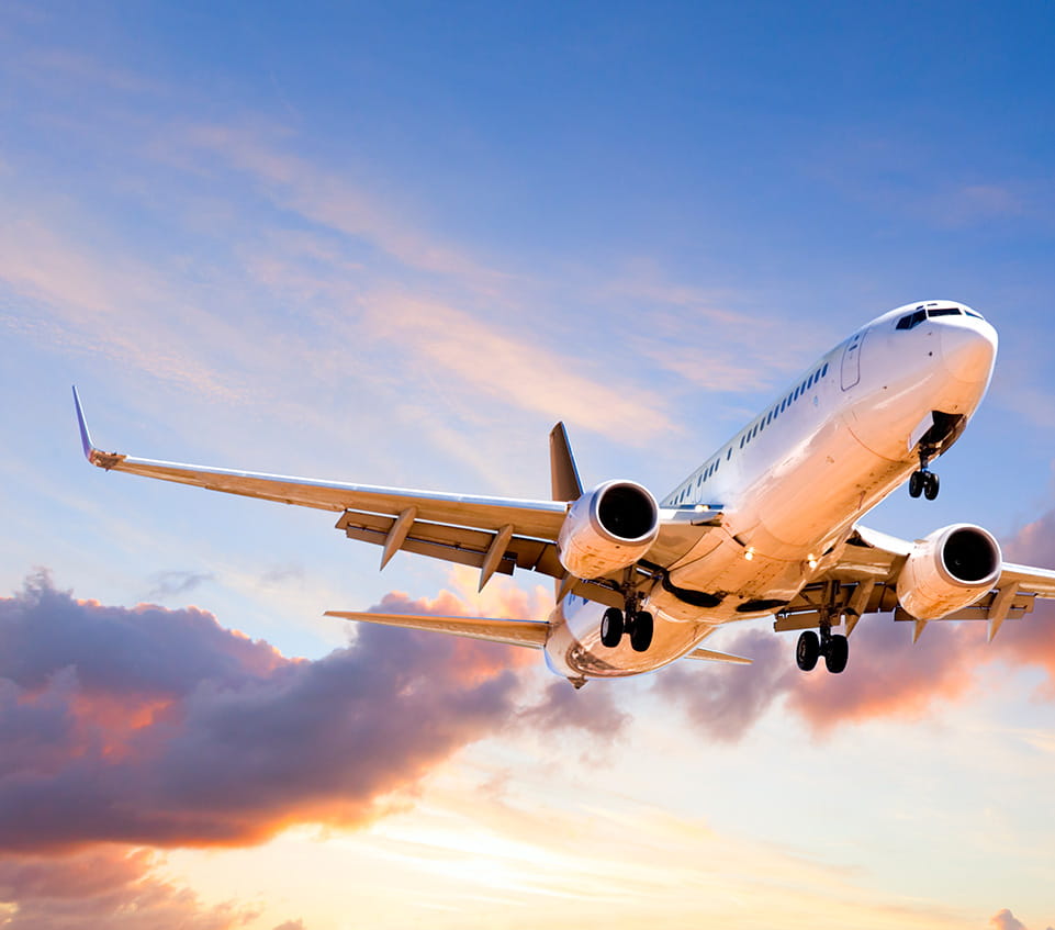 Twin-engine commercial jet with winglets and deployed landing gear descending against cloud-filled sky at sunset.