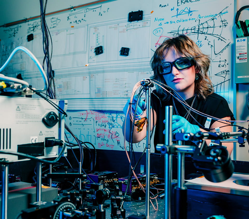 Student working with biomedical instrumentation and tubing setup in engineering lab, wearing protective glasses and gloves, with circuit diagrams and equations on whiteboard behind.