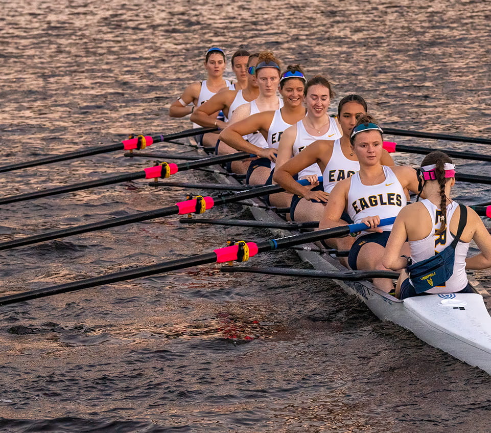 Women’s rowing team in eight-person shell with coxswain steering, athletes in Embry-Riddle Eagles uniforms rowing in sync on water.