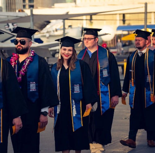 Graduates in caps and gowns walking on airfield with small aircraft parked in background.
