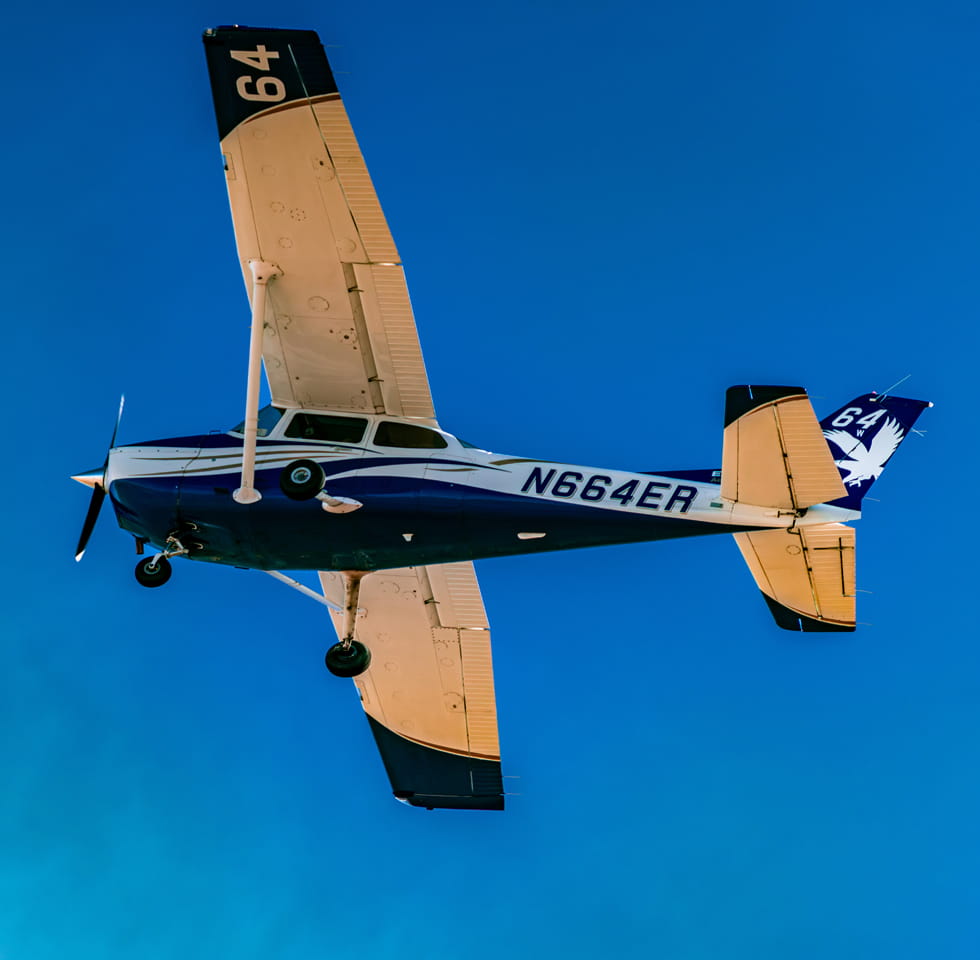 A small blue-and-white propeller airplane flying overhead against a clear blue sky.