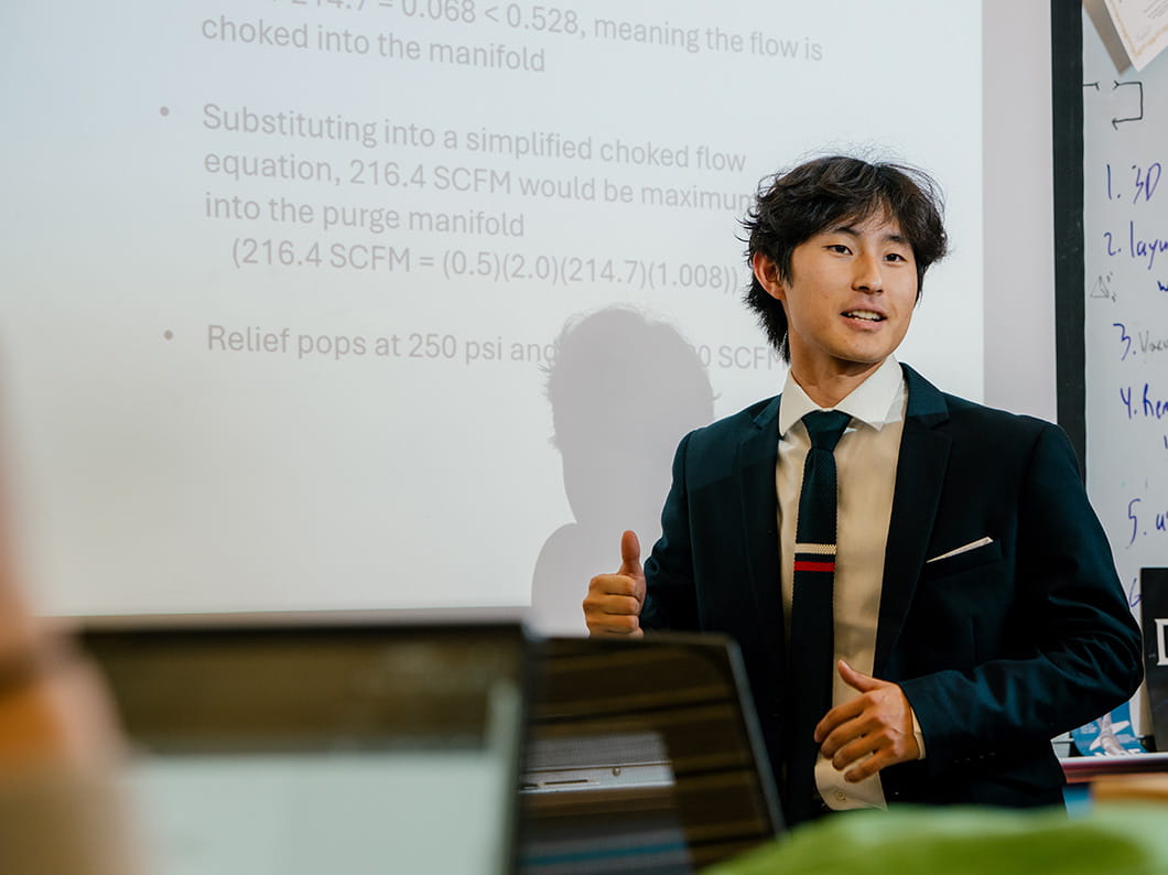 Student in business attire presenting fluid flow calculations on projected slide, including SCFM values and choked flow equation, gesturing with hand while speaking in classroom with whiteboard at side.
