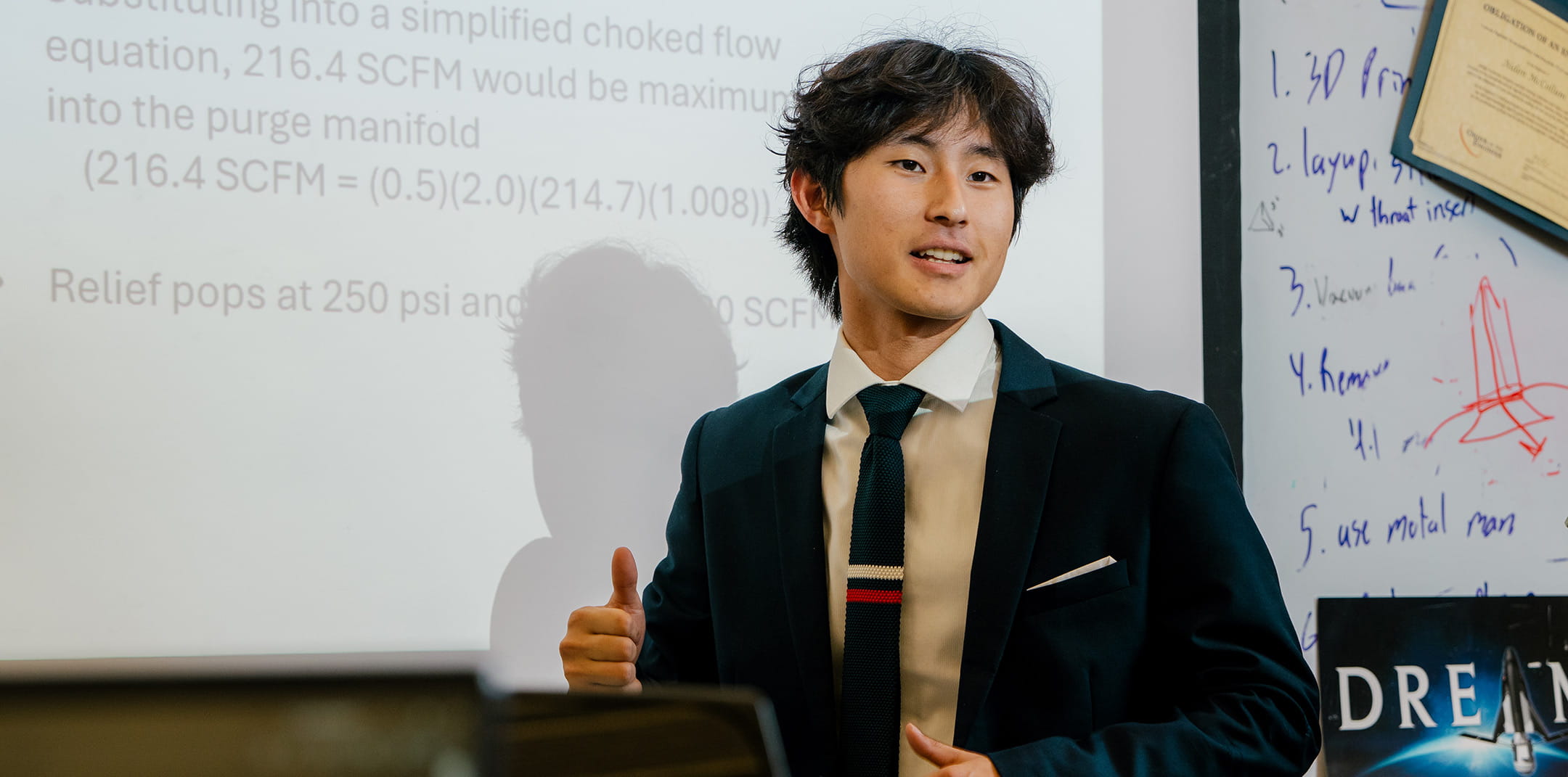 Student in business attire presenting fluid flow calculations on projected slide, including SCFM values and choked flow equation, gesturing with hand while speaking in classroom with whiteboard at side.