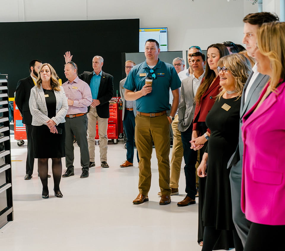 Members of advisory board and staff standing in an aerospace lab with tool cabinets and equipment, wearing business attire and listening.