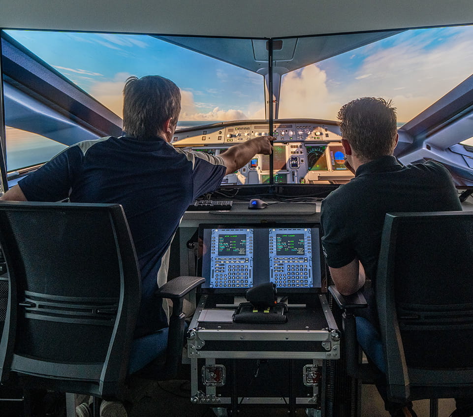 Two students operating advanced flight simulator with dual flight management system keypads and glass cockpit displays, one pointing at primary flight display while viewing horizon on wraparound screen.