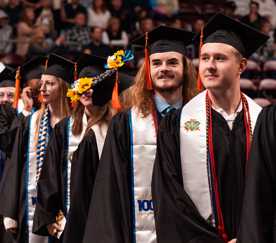 Graduating students in caps and gowns wearing honor cords and stoles, including decorated mortarboard with sunflowers, standing in line during commencement ceremony with audience in background.