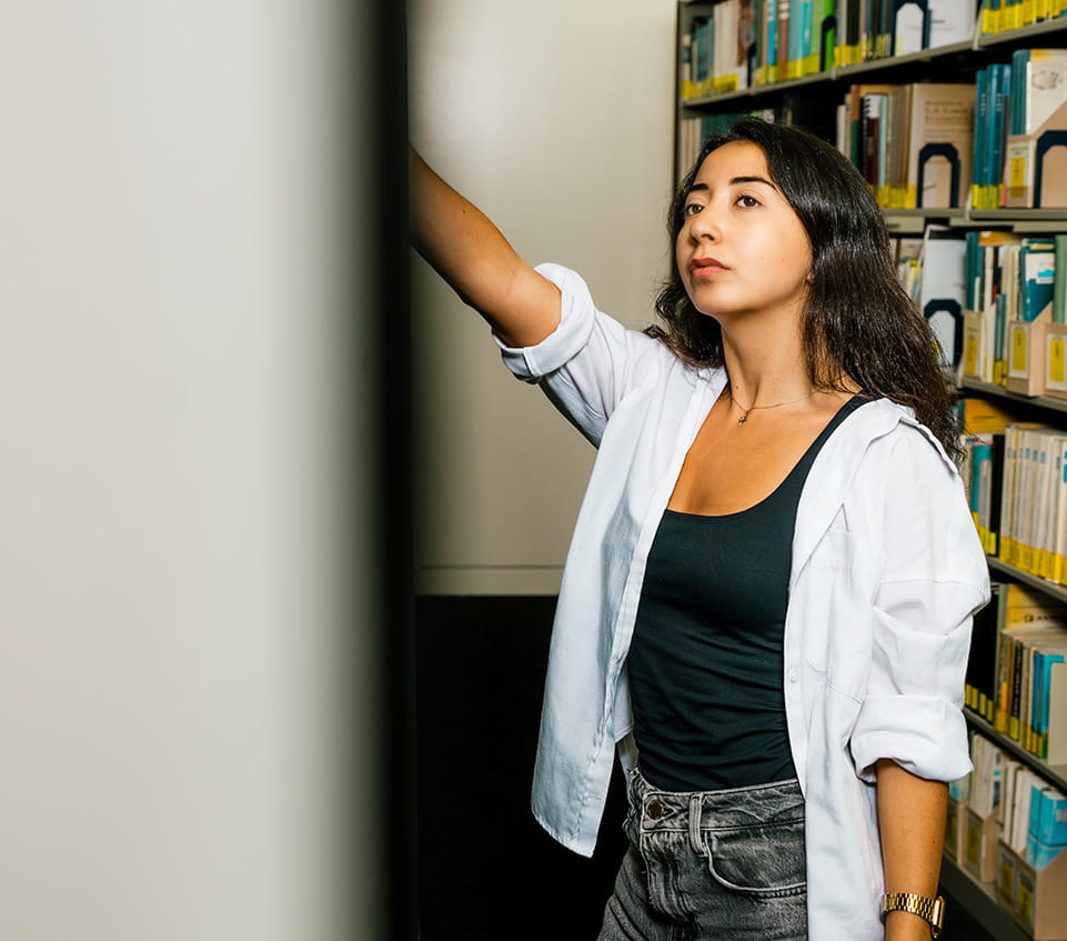 Student reaching for book on library shelf, standing in an aisle of campus library stacks.