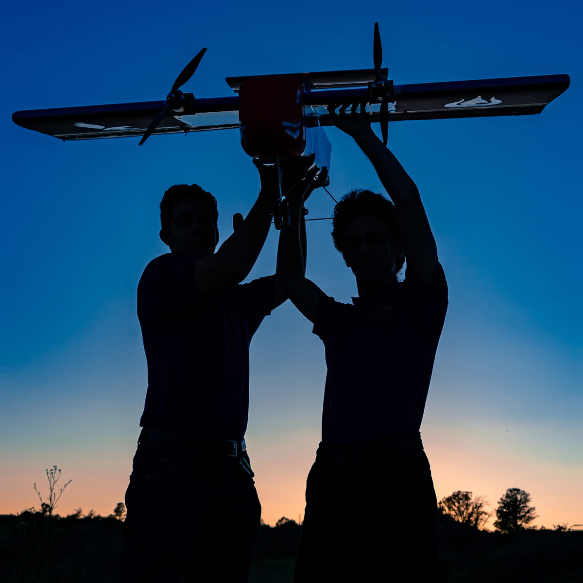 Students launching small fixed-wing unmanned aircraft by hand at dusk with sky gradient in background.
