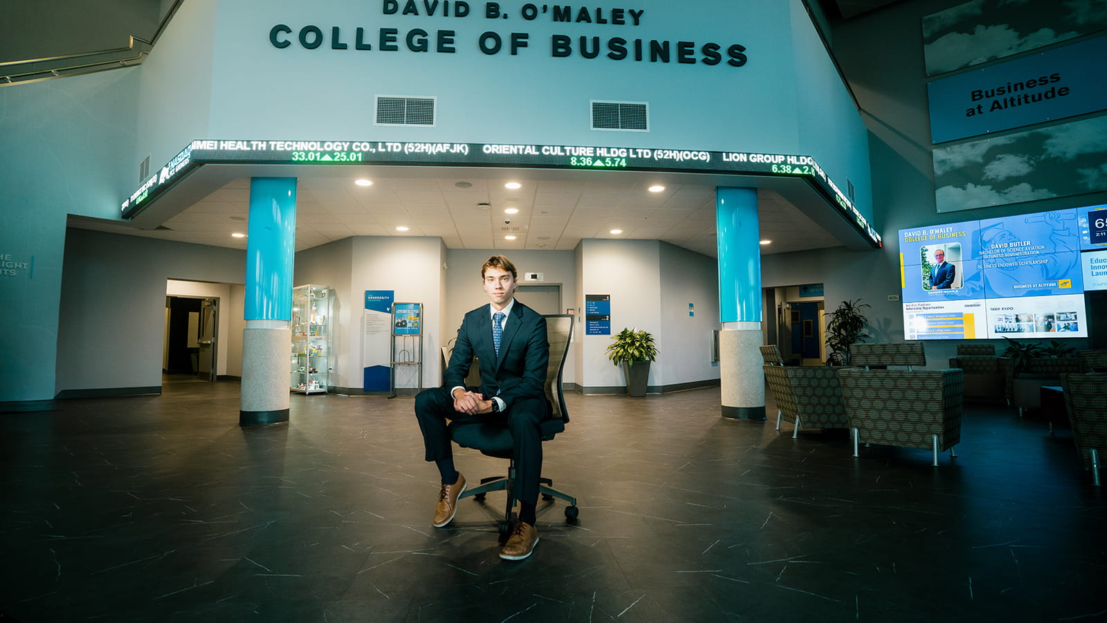 Embry-Riddle student sits on a computer chair in the lobby of the College of Business as a stock ticker scrolls on the wall behind him