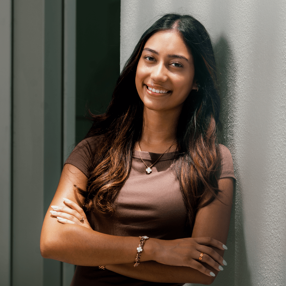 Alisha leans against a wall with arms crossed smiling.