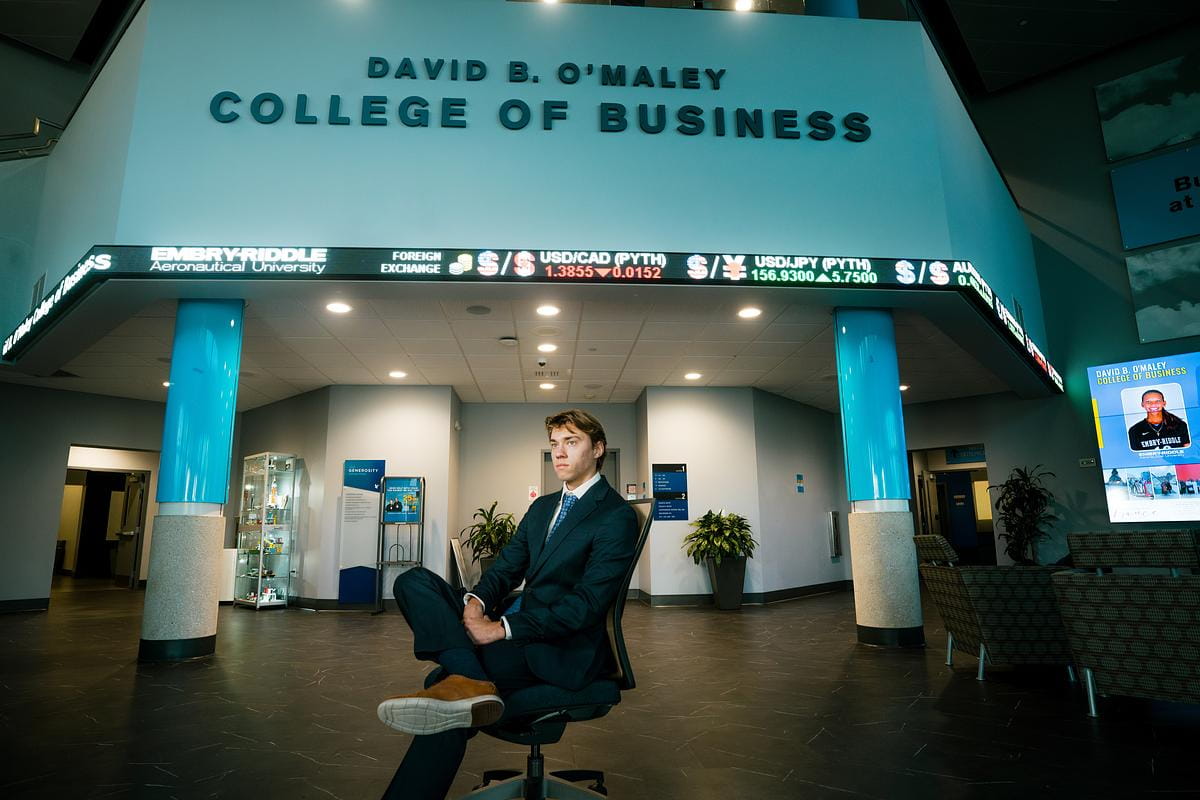 Student in David B. O'Maley sits in the lobby with a stock ticker screen behind him.