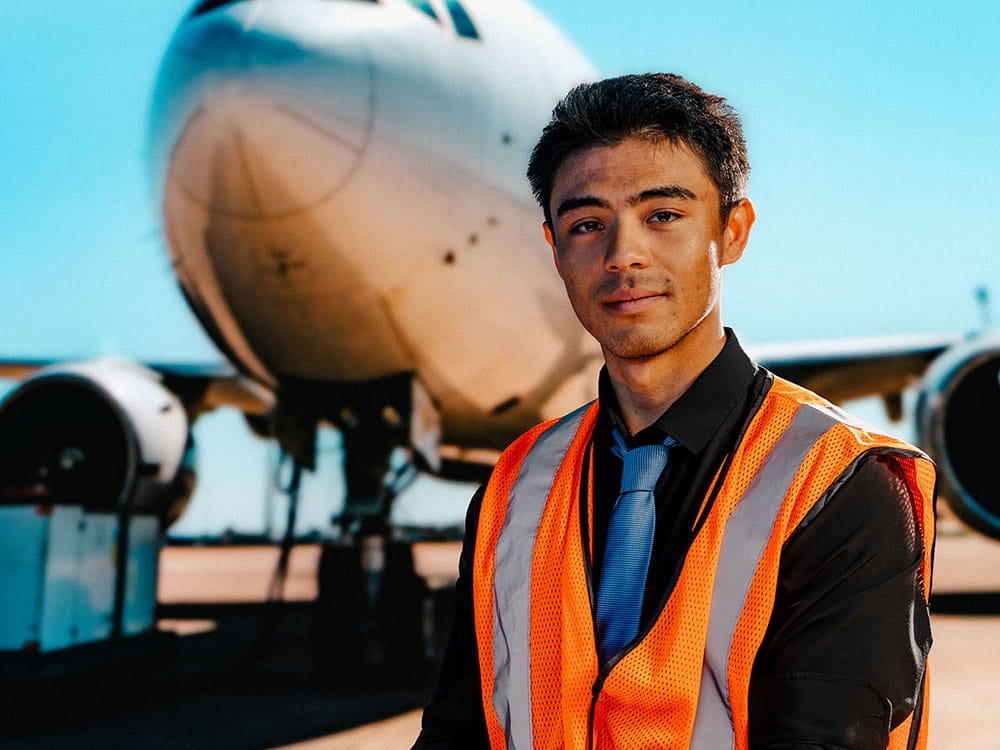A student wearing a safety vest stands in front of an airplane on a tarmac