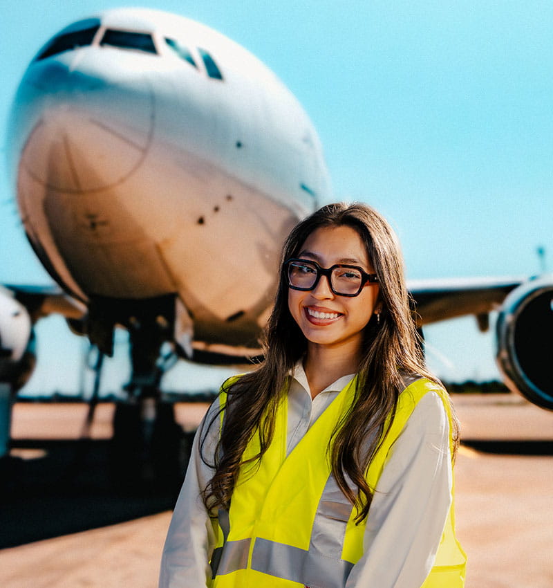 A student wearing a safety vest stands in front of a plane on the tarmac