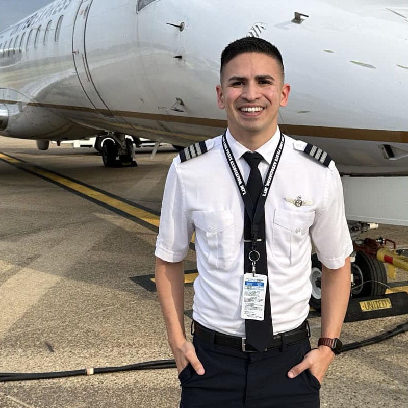 Juan Pena wears a pilot uniform and stands next to a plane on the tarmac