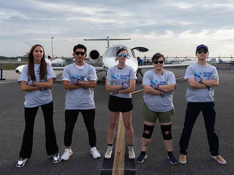 Five Embry-Riddle students wearing Aviation Week shirts stand in front of a plane with their arms crossed.