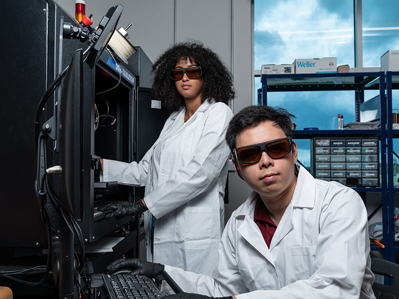 Two students wear white lab coats while working in an electrical engineering lab